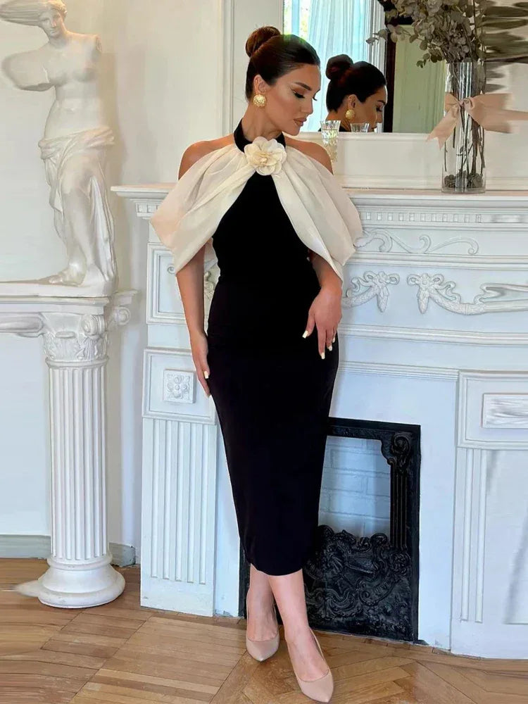 Woman wearing a black sheath dress with a white sheer off-the-shoulder overlay adorned with a large flower accent, standing in a formal setting.