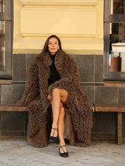 Woman wearing a long, fluffy brown faux fur coat over a dark skirt and black shoes, sitting on a wooden bench.