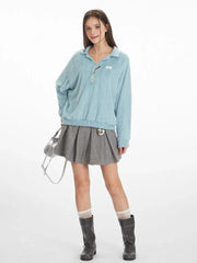 Full body shot of a model wearing a light blue fuzzy knit collared v-neck sweater with a grey pleated mini skirt, white and grey striped socks, and black boots, holding a silver handbag.