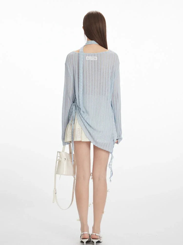 back view of a model wearing a light blue sheer knit sweater with an asymmetrical hem and a neck scarf, paired with a white lace skirt and white heels, holding a white handbag