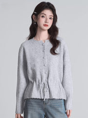 A woman stands wearing a light grey knit cardigan with a drawstring waist and button front closure, paired with blue jeans.