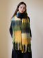Woman wearing a long, oversized scarf with a green, yellow, and black plaid pattern and fringe detail.