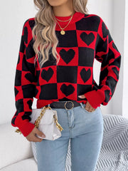 Woman wearing a red and black checkerboard heart print knitted sweater with light blue jeans and a white handbag, holding her hand on her hip