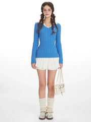 Woman wearing a blue cable knit V-neck sweater with a white collar over a white pleated skirt, cream leg warmers, and a cream handbag, front view.