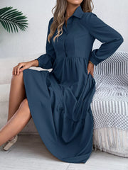Woman sitting on a couch wearing a long-sleeved blue midi dress with a collared neckline and tiered hem