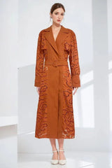 Model wearing a rust-colored laser-cut floral trench coat with a belted waist and notched lapels.