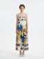 Woman wearing a white maxi dress with a colorful abstract print, spaghetti straps, and white heels, standing against a white background.