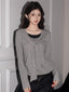 Woman wearing a grey long-sleeved top with a draped overlay and black pants, standing against a wall.
