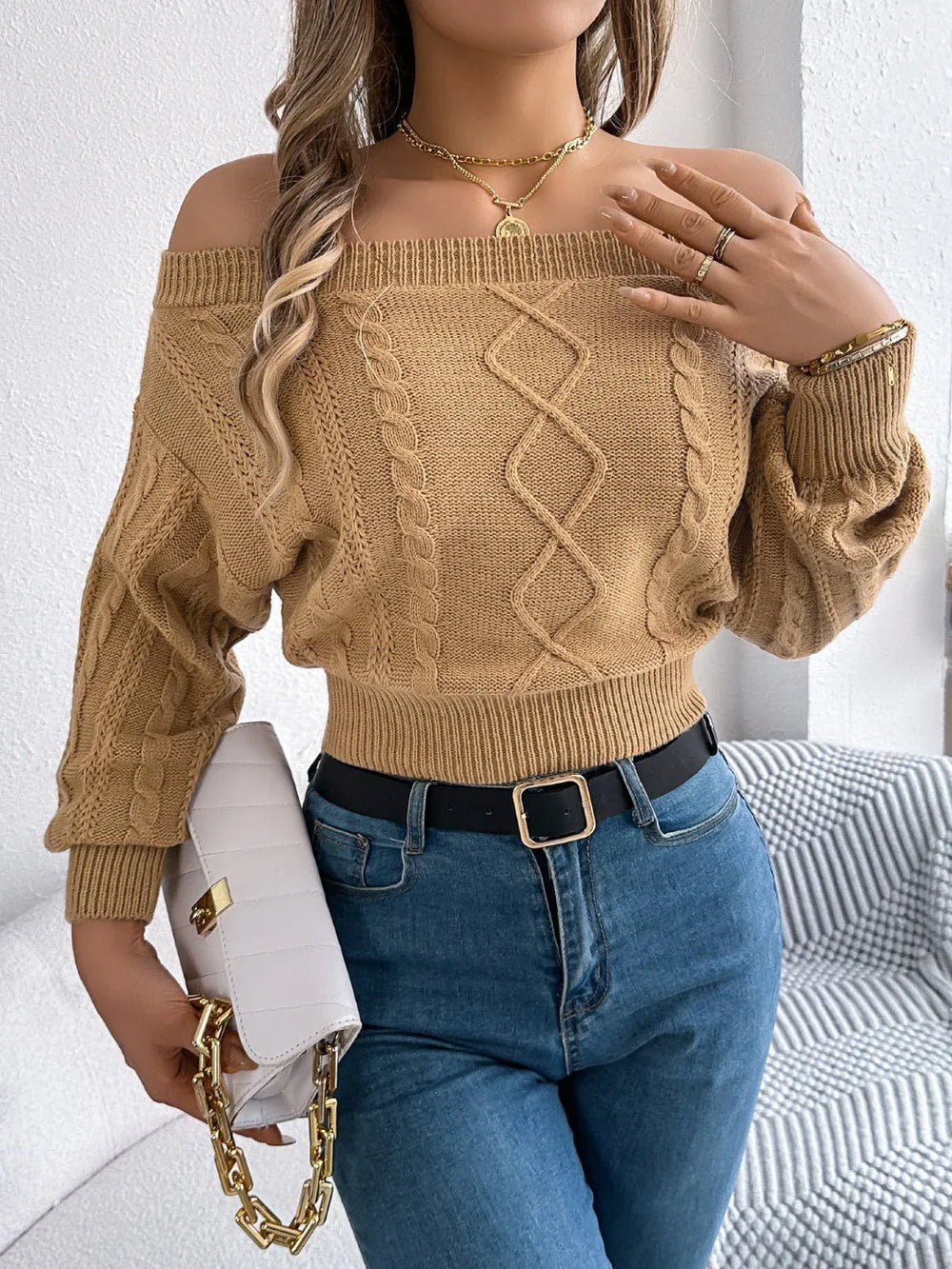 Woman in a tan off-the-shoulder cable knit cropped sweater, blue jeans, and a black belt, holding a white handbag with a gold chain.