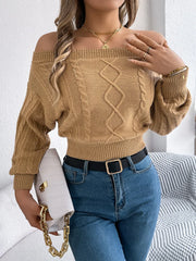 Woman in a tan off-the-shoulder cable knit cropped sweater, blue jeans, and a black belt, holding a white handbag with a gold chain.