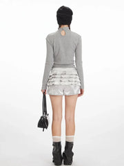 Back view of a model wearing a grey long-sleeved top with a graphic print and silver ruffled shorts, black beanie, white socks, and black boots.