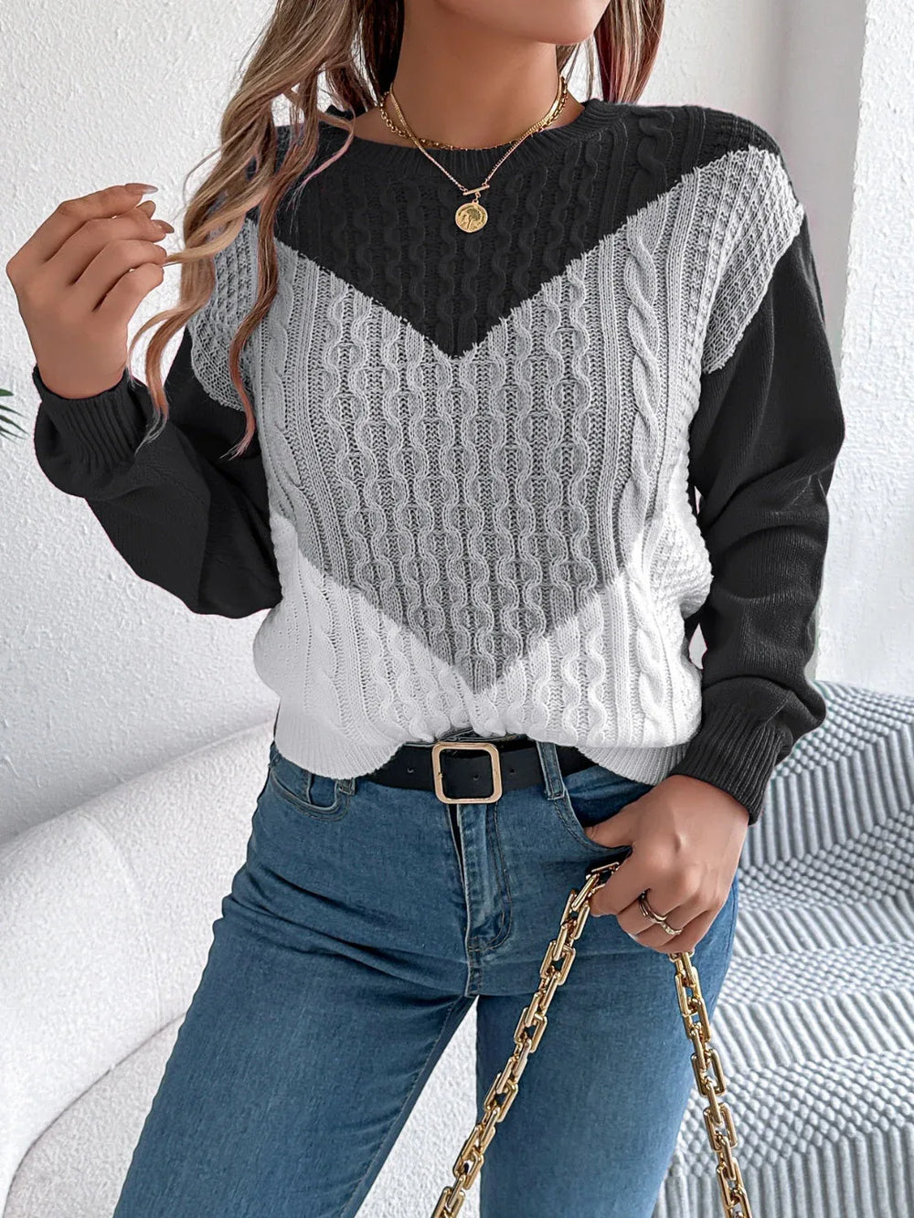 Woman wearing a black, grey, and white color block chevron cable knit sweater and blue denim jeans, accessorized with a gold chain necklace and a black belt.