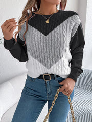 Woman wearing a black, grey, and white color block chevron cable knit sweater and blue denim jeans, accessorized with a gold chain necklace and a black belt.