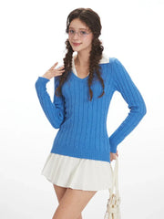 Close-up of a woman wearing a blue cable knit V-neck sweater with a white collar and a white pleated skirt, posing with her hand on her hip, front view.