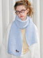 A young woman with curly hair and glasses wears a light blue fuzzy knit scarf with an embroidered dog detail.
