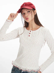 Model posing with a red baseball cap and a white long-sleeve top with a floral print and red trim.