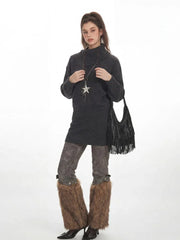 Full-body shot of a woman in a charcoal grey oversized turtleneck knit tunic, patterned leggings, and brown furry leg warmers, accessorized with a star pendant necklace and a black fringed handbag.