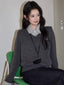 Woman sitting on a green chair wearing a grey v-neck sweater over a checkered collared shirt, dark wash wide leg jeans, and a black crossbody pouch.