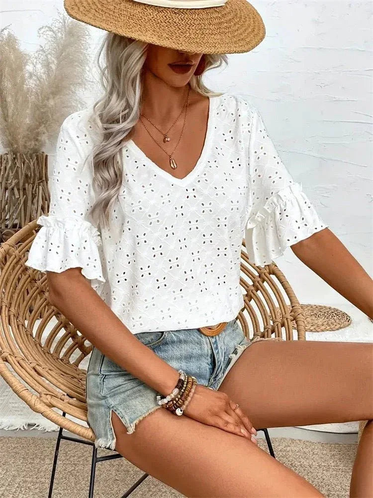 A woman sits in a rattan chair wearing a white eyelet top with ruffled sleeves and denim shorts, accessorized with a straw hat.