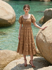 Woman wearing a brown puff-sleeve midi dress with a smocked bodice and tiered skirt featuring a sun and geometric print, leaning on a rock on a beach with the ocean in the background.