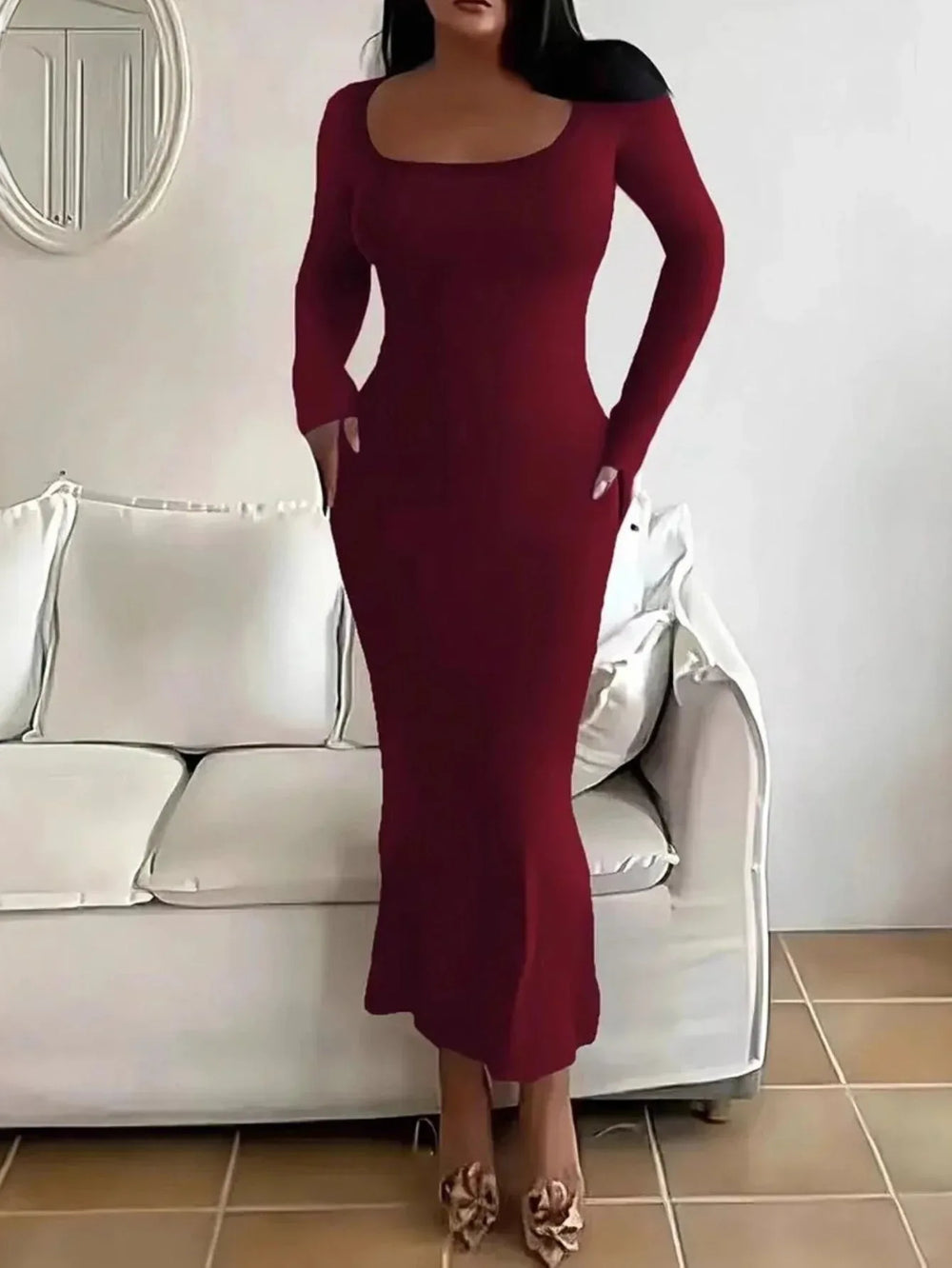 Woman wearing a long-sleeved, scoop-neck, form-fitting, floor-length mermaid silhouette dress in dark red, standing in a living room.