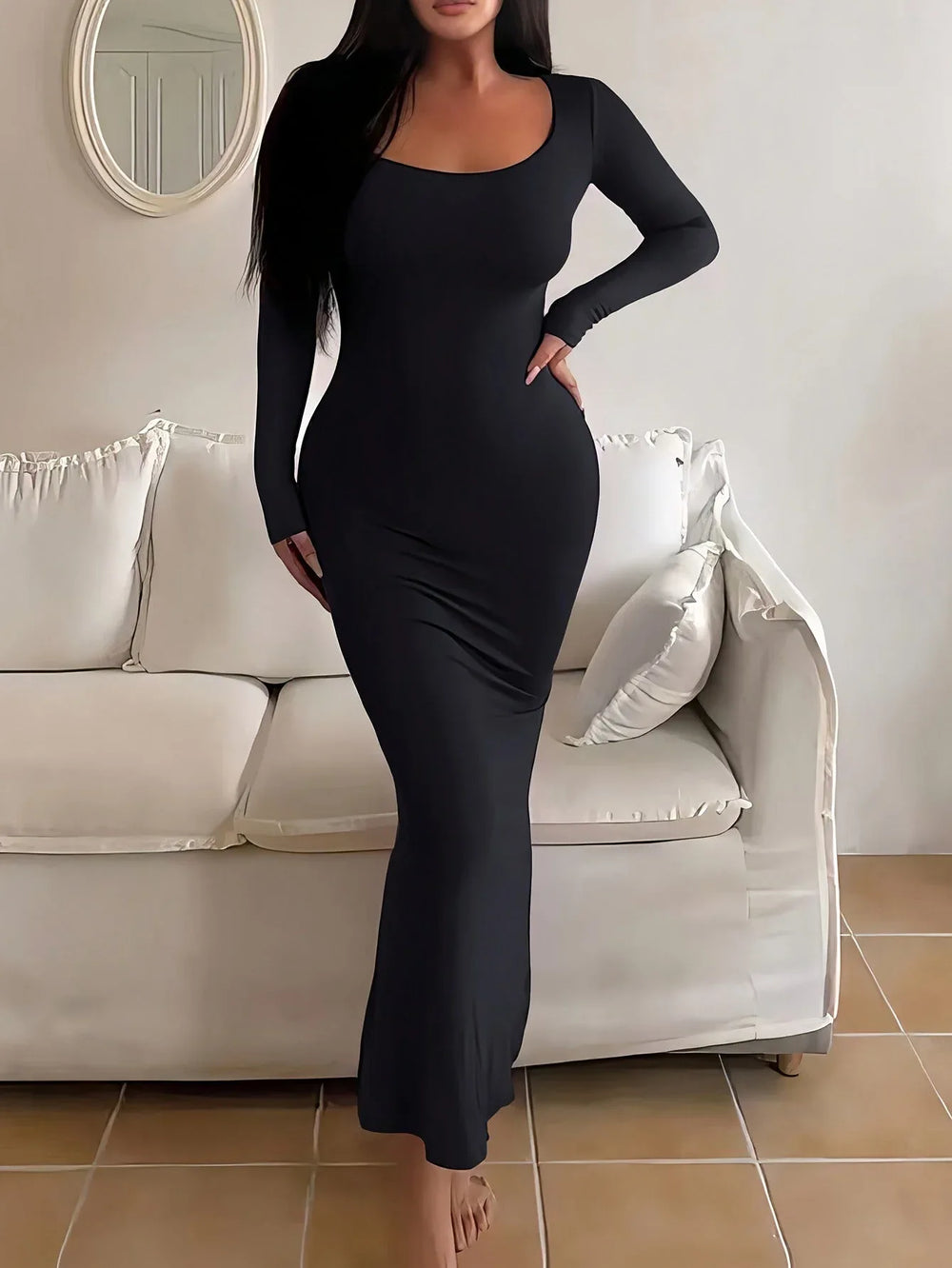 Woman wearing a long-sleeved, scoop-neck, form-fitting, floor-length mermaid silhouette dress in black, standing in a living room.