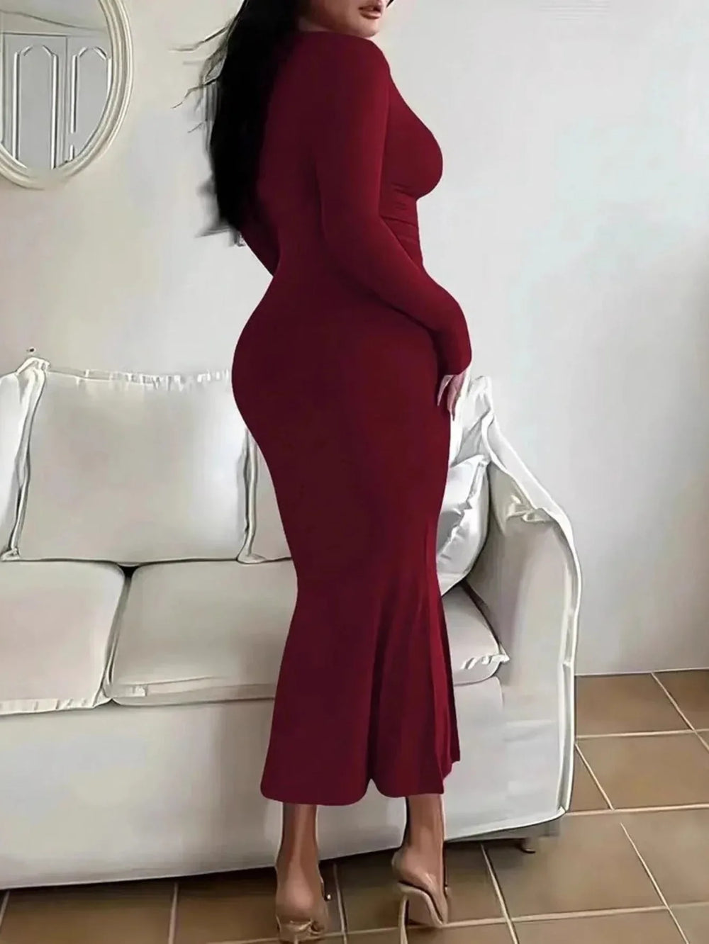 Back view of a woman wearing a long-sleeved, scoop-neck, form-fitting, floor-length mermaid silhouette dress in dark red, standing in a living room.