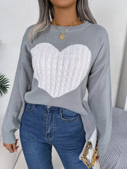 Close-up of a woman wearing a grey knit sweater with a white cable knit heart graphic, blue jeans, and gold necklaces.