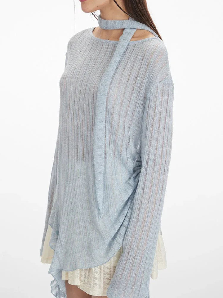 close-up side view of a model wearing a light blue sheer knit sweater with an asymmetrical hem and a neck scarf, paired with a white lace skirt and white heels, holding a white handbag