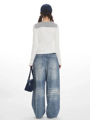 Back view of a model wearing a white long-sleeved top with striped trim and wide-leg blue jeans, holding a navy handbag and wearing a navy baseball cap.