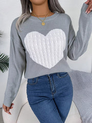 Woman wearing a grey knit sweater with a white cable knit heart graphic, blue jeans, and gold necklaces.