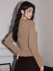 Back view of a woman wearing a tan ribbed knit long-sleeve top with a tie-front closure and ruffled hem, paired with a brown pleated skirt.