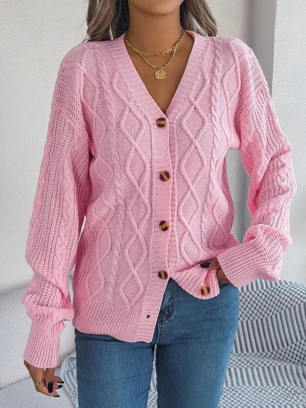 front view of a woman wearing a pink cable knit cardigan with a v-neckline and button-down closure, paired with blue jeans