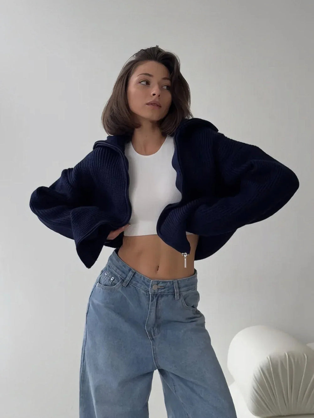 Woman wearing a white crop top and blue wide-leg jeans, with a navy blue zip-up cardigan open.