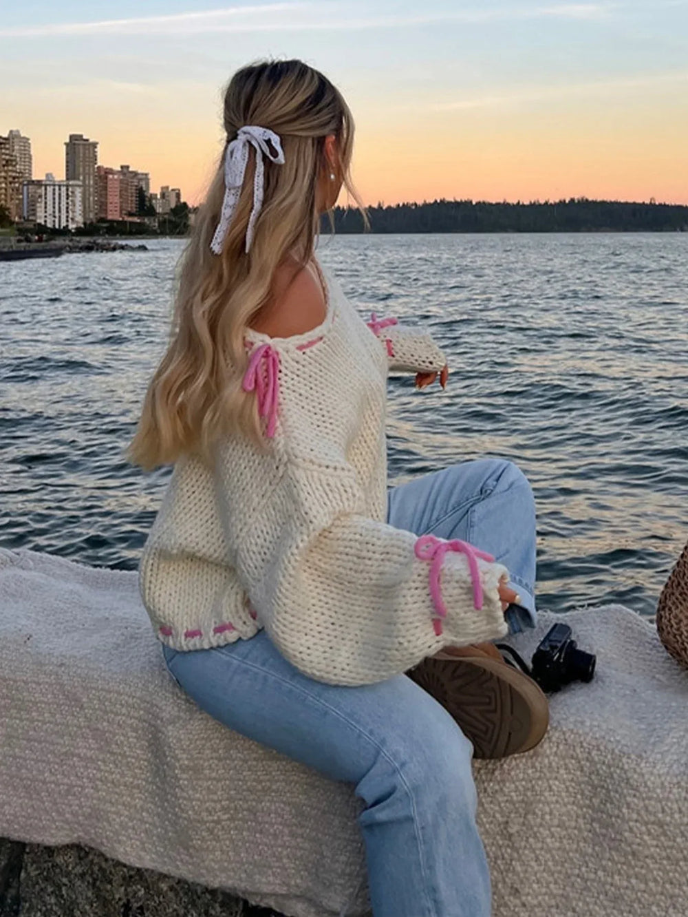 Woman with blonde hair wearing a cream chunky knit sweater with pink tie details, sitting by the water at sunset.
