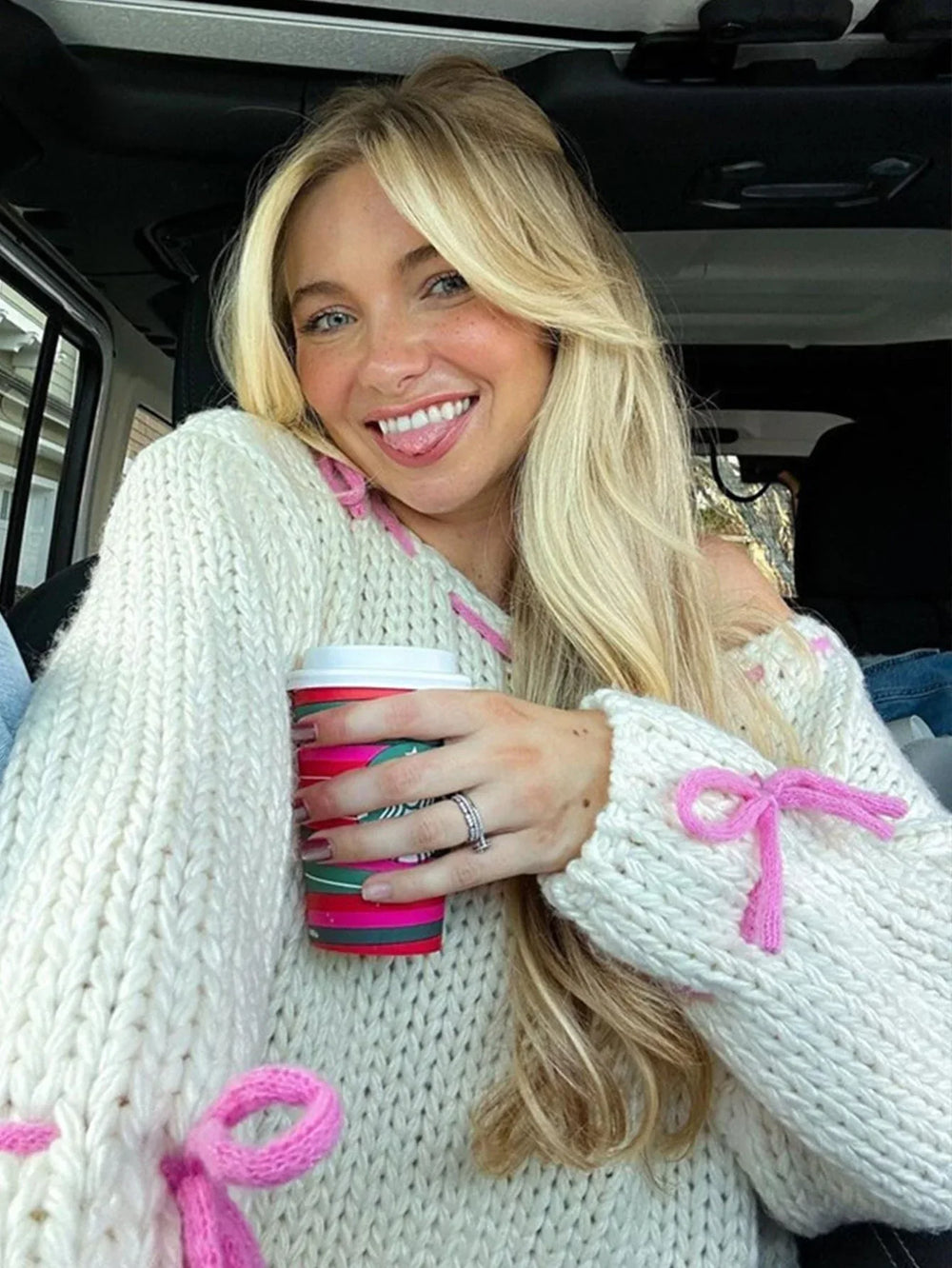 Woman with blonde hair wearing a cream chunky knit sweater with pink tie details, holding a coffee cup and smiling.