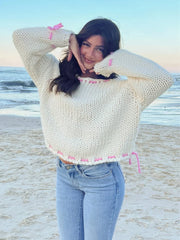 Woman with dark hair wearing a cream chunky knit sweater with pink stitching, posing on a beach.