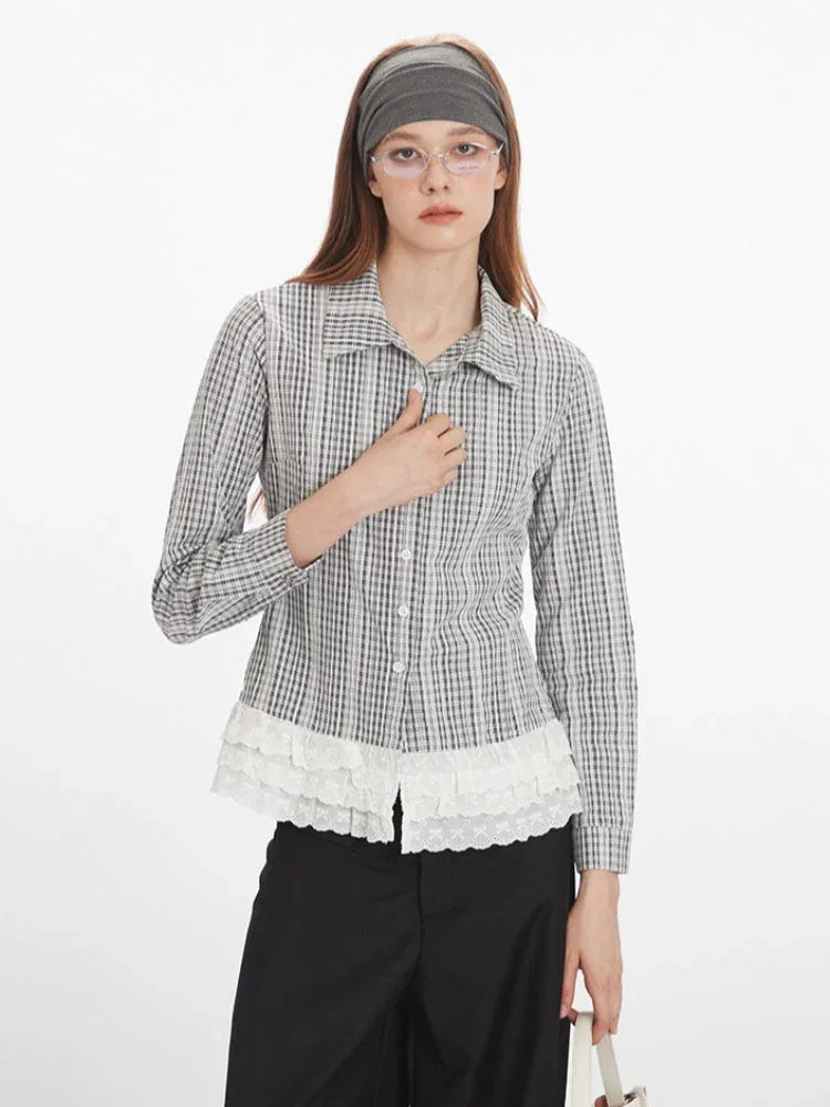Front close-up of a woman wearing a long-sleeved plaid collared shirt with a white ruffled hem and black pants, with one hand on her chest.