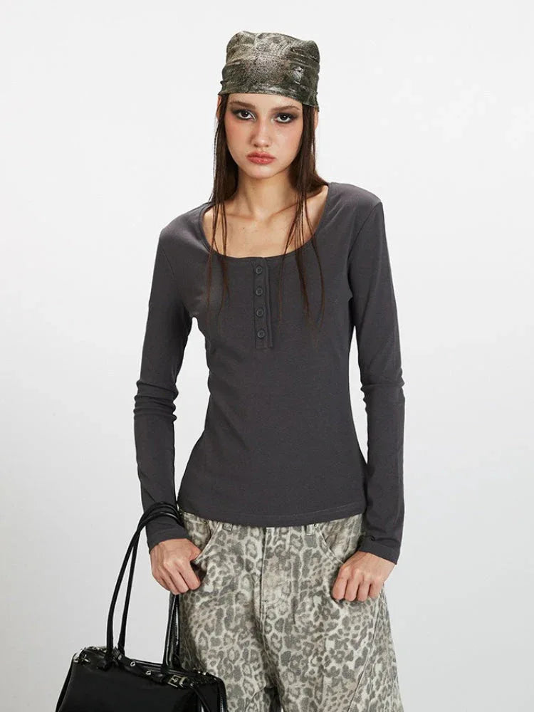 Close-up front view of a woman wearing a dark grey long-sleeve top with a square neckline and button details, paired with wide-leg grey leopard print pants. She is holding a dark handbag and wearing a patterned headscarf.
