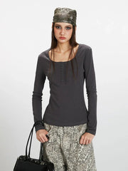 Close-up front view of a woman wearing a dark grey long-sleeve top with a square neckline and button details, paired with wide-leg grey leopard print pants. She is holding a dark handbag and wearing a patterned headscarf.