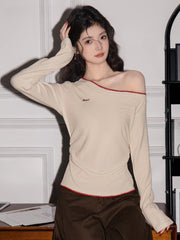 Woman wearing a cream off-the-shoulder long-sleeve top with red trim and brown pants, touching her hair.
