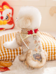 The back view of a small white dog wearing a gold and floral brocade coat with faux fur trim and pearl accents.