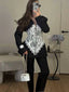 Woman wearing a black blazer with a white lace corset overlay, black leggings, and black boots, holding a white handbag.