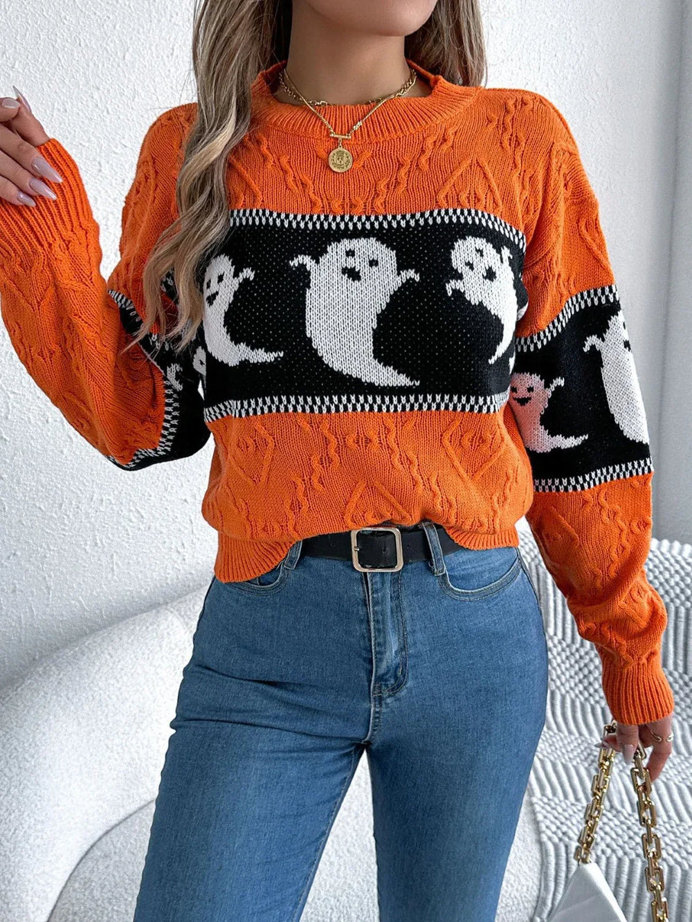 Woman wearing an orange Halloween sweater with a ghost print and blue jeans, holding a white purse.
