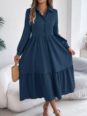 Woman standing holding a straw bag and wearing a long-sleeved blue midi dress with a collared neckline and tiered hem