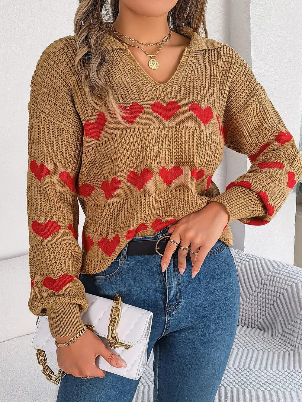 Woman holding a white clutch and wearing a tan knit sweater with red heart pattern and blue jeans