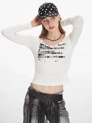 Close-up front view of a woman wearing a white long-sleeved graphic t-shirt with a scoop neck and studded details, grey distressed jeans with a black fringed belt, and a black beaded necklace.