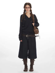 Woman wearing a dark long-sleeved V-neck top, dark wide-leg cropped denim pants, a brown leather belt, brown boots, and a brown leather crossbody bag, posing against a white background.