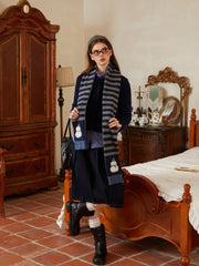 Woman wearing a navy blue sweater, a blue and white checkered shirt, a dark navy pleated skirt, white socks, black boots, and a grey and navy striped scarf with snowman embellishments. She is standing in a bedroom with a wooden wardrobe and a bed.