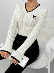 Woman sitting and holding a book, wearing a white cable knit cropped V-neck sweater with black trim and a black bow detail, paired with black pants.
